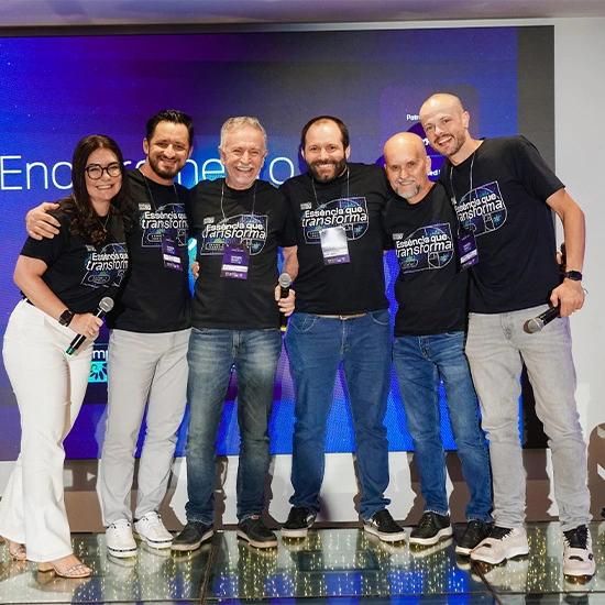 Equipe Seprol reunida no palco de um evento, posando com microfones, vestindo camisetas 'Essência que transforma'
