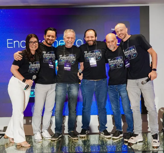 Equipe Seprol reunida no palco de um evento, posando com microfones, vestindo camisetas 'Essência que transforma'